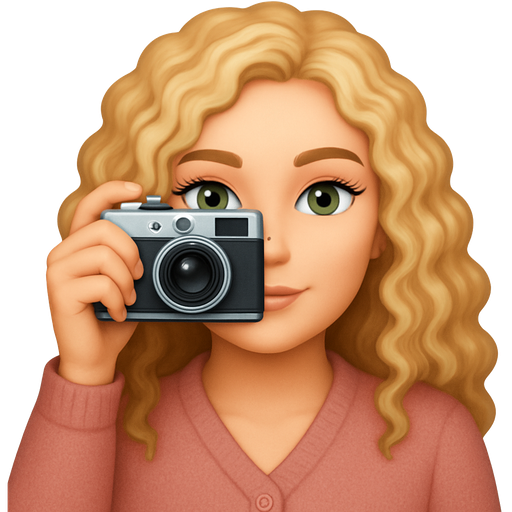 Woman with long light blonde curly hair and fair skin and green eyes and dark eyelashes, wearing a soft rose cardigan, holding a film camera up to her right eye.